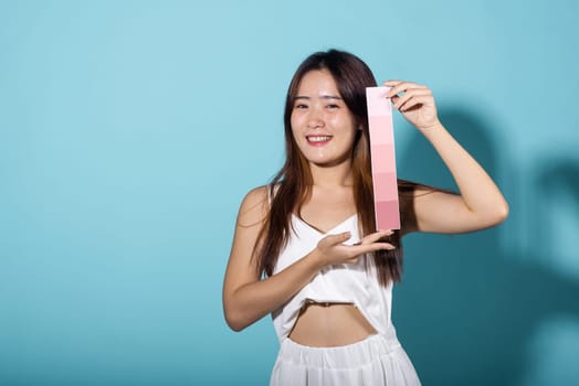 Portrait of happy female hold color card under sun protection studio shot isolated on blue background, Asian beautiful young woman smiling and holding skin color scale paper, skincare beauty face