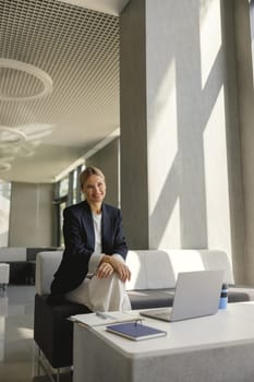 Pretty female entrepreneur working on laptop sitting in modern office hall and looks camera
