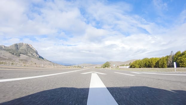 On a crisp winter day, a car cruises along the iconic Highway 101 near San Luis Obispo, California. The surrounding landscape is brownish and subdued, with rolling hills and patches of coastal vegetation flanking the winding road.