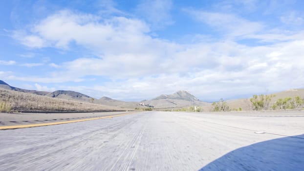 On a crisp winter day, a car cruises along the iconic Highway 101 near San Luis Obispo, California. The surrounding landscape is brownish and subdued, with rolling hills and patches of coastal vegetation flanking the winding road.