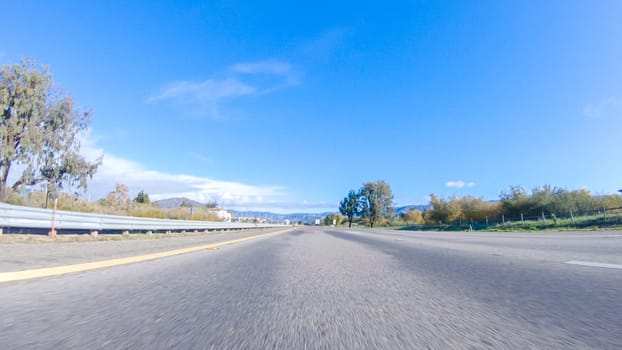 On a crisp winter day, a car cruises along the iconic Highway 1 near San Luis Obispo, California. The surrounding landscape is brownish and subdued, with rolling hills and patches of coastal vegetation flanking the winding road.