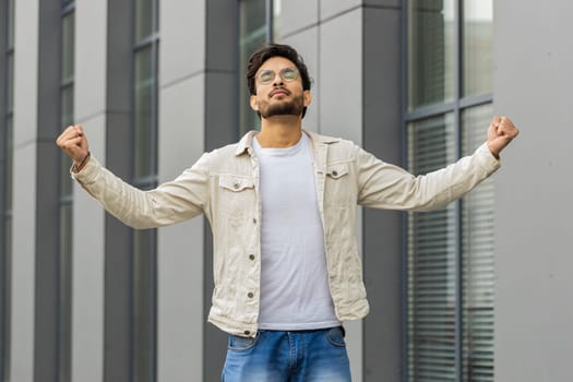 Young Indian man taking a deep breath of fresh air, relaxing, taking a break, resting, meditating, calm down outdoors. Arabian Hindu guy enjoying happy day in downtown city street. Business people