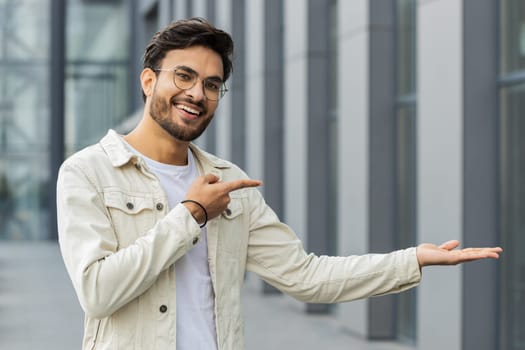 Happy Indian man showing pointing empty place advertising area for commercial text copy-space for goods promotion sale discount outdoors. Arabian young guy in downtown city street. Business people