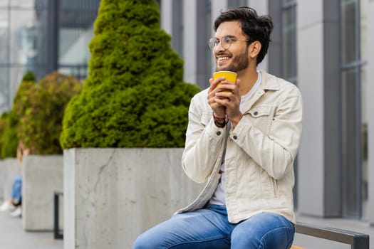 Happy Indian man enjoying morning coffee hot drink and smiling outdoors. Relaxing, taking a break. Arabian guy in city downtown street sitting on bench, drinking coffee to go. Town lifestyles outside