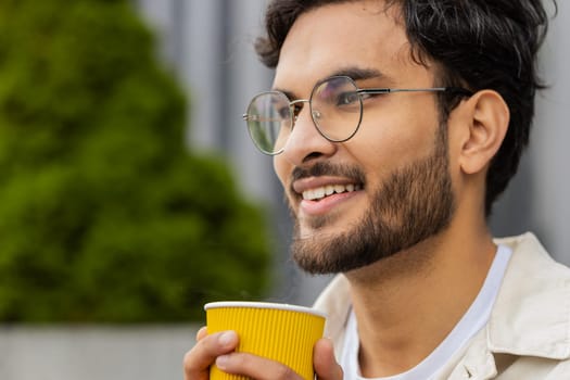 Close-up happy Indian man enjoying morning coffee hot drink and smiling outdoors. Relaxing, taking a break. Arabian Hindu guy in city downtown street, drinking coffee to go. Town lifestyles outside