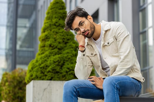 Tired sad Indian man feeling worried about financial problem stress outdoors. Arabian Hindu guy frustrated thinking of money debt budget loss bankruptcy sitting on downtown street bench. Overworking