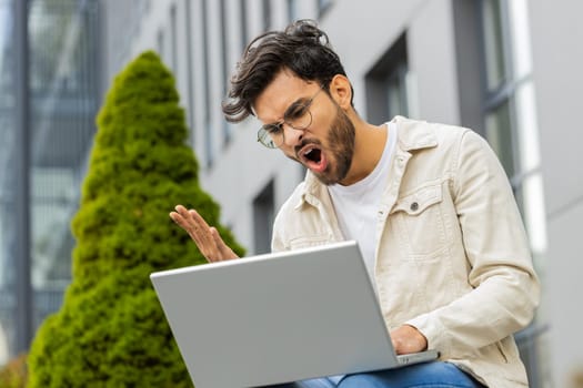 Displeased Indian man use laptop typing browsing working, loses becoming surprised sudden lottery results bad news fortune loss game fail computer virus. Arabian Hindu guy sitting on street city bench