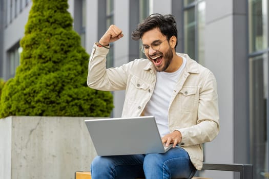 Overjoyed happy young Indian man working on laptop scream in delight raise hands in triumph winner gesture celebrate success win money outdoors. Arabian Hindu guy sitting on downtown street bench