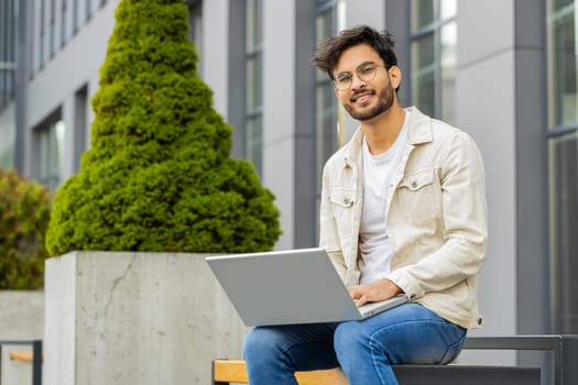 Happy Indian man freelancer working on laptop computer sends messages, makes online purchases, watching movies, reading email outdoors. Arabian Hindu guy looking at camera sitting on street bench