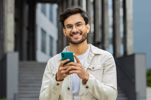 Young smiling Indian man using smartphone typing text messages, reading email, browsing news outdoors. Arabian Hindu guy use mobile social media app looking at camera standing in downtown city street