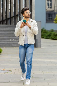 Young smiling Indian man using smartphone typing text messages, reading email, browsing news outdoors. Arabian Hindu guy using mobile social media app walking in downtown city street. Business people