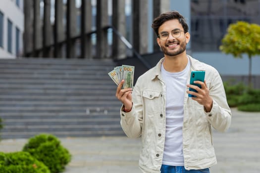 Happy rich champion Indian man counting money dollar cash, use smartphone calculator app plans to order gifts and food delivery online, booking. Arabian Hindu guy in city street outdoors. Copy-space