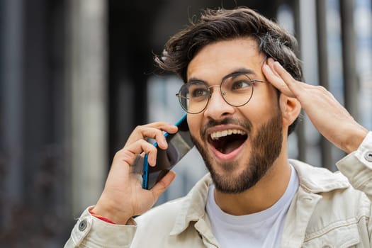 Phone call, good news, gossip. Happy surprised Indian man in pleasant conversation on smartphone, enjoying talking with friend outdoors. Arabian excited guy standing in city street. Town lifestyles