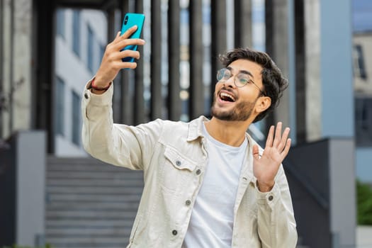 Young Indian man blogger taking selfie on smartphone, communicating video call online with subscribers recording stories for social media vlog outdoors. Arabian Hindu guy standing in urban city street