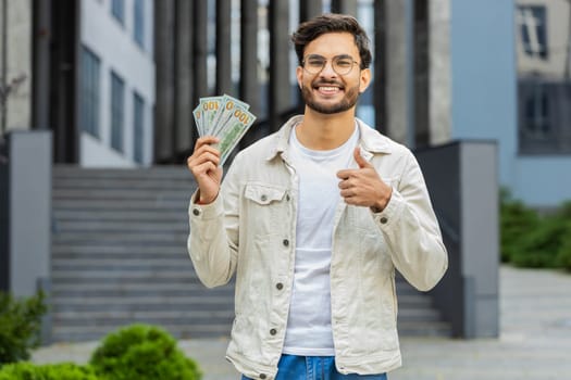 Happy Indian man counting money cash holding dollars banknotes, bills budget count salary profit reward bribe outdoors. Successful rich Arabian Hindu guy showing thumbs up in downtown city street