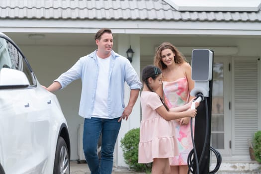 Happy little young girl learn about eco-friendly and energy sustainability as she help her family recharge electric vehicle from home EV charging station. EV car and modern family concept. Synchronos