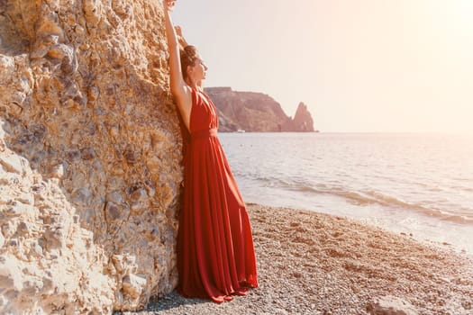 Side view a Young beautiful sensual woman in a red long dress posing on a rock high above the sea during sunrise. Girl on the nature on blue sky background. Fashion photo.