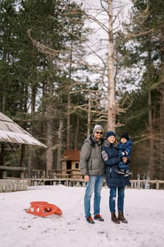 Smiling dad with sled stands hugging mom with a little boy in her arms in a snowy forest. High quality photo