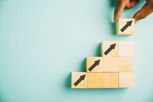 Male hands arranging stacked wooden blocks as steps on a white table. Business growth concept with an arrow pointing upwards, symbolizing future development and achieving goals. Copy space available.