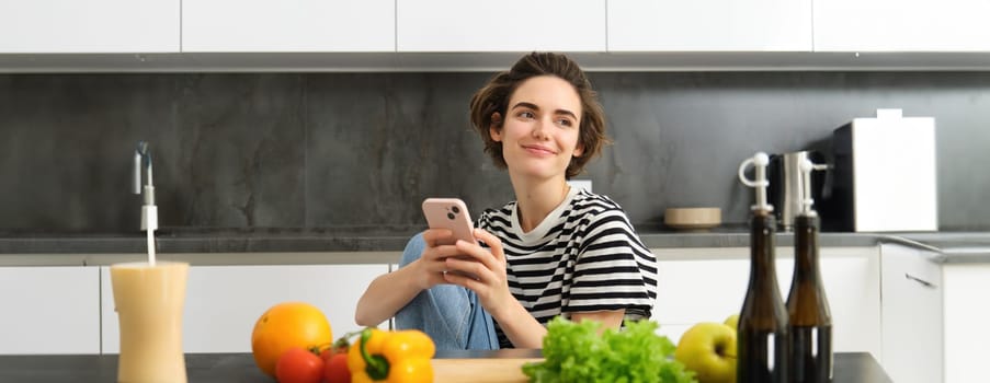 Portrait of brunette young woman with smartphone, sitting in the kitchen, watching cooking recipe videos, has chopping board and vegetables.