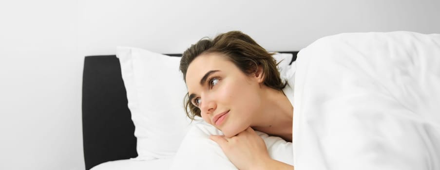 Close up portrait of beautiful brunette woman, waking up in her bed, looking at alarm clock, staring aside with smiling dreamy face.