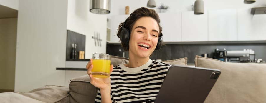 Portrait of woman in headphones, sitting on sofa with tablet and drinking orange juice, watching tv show on her gadget, using social media application, spending time at home.