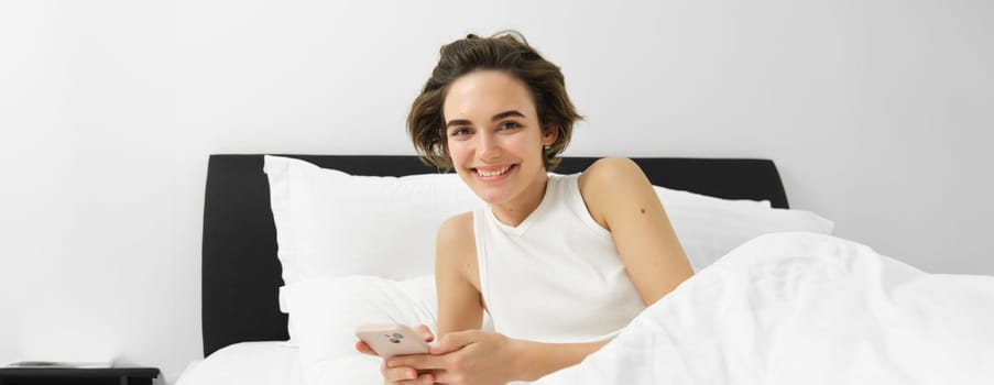 Image of woman checking her sleep app on mobile phone, lying in bed and smiling, using smartphone in bedroom.