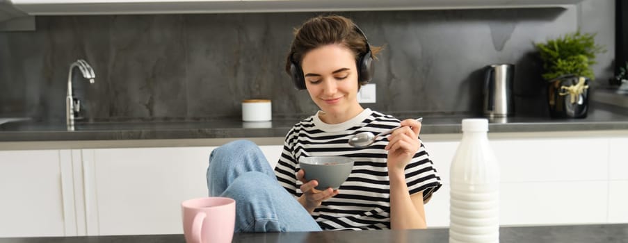 Portrait of smiling young candid woman, student eating morning cereals, having her breakfast, listening music in wireless headphones, sitting in the kitchen.