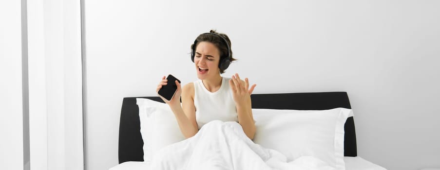 Carefree woman in bed, wakes up and listens to music in wireless headphones, dancing and singing, using mobile phone as microphone.