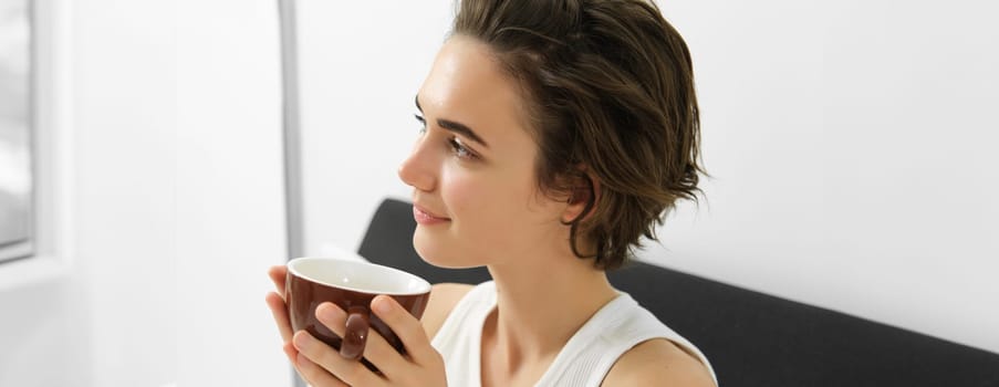 Close up portrait of relaxed woman, resting in bed, waking up and drinking morning cup of coffee, smiling with happy, pleased face.