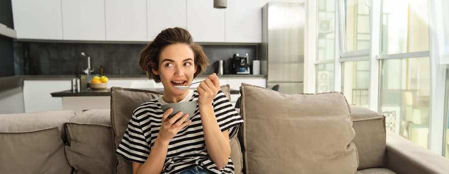 Portrait of cute young woman eating breakfast, having cereals with milk on sofa in living room, smiling and looking satisfied.