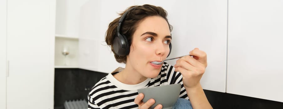 Cute young woman in headphones, eating with spoon from bowl, having cereals for breakfast, sitting in kitchen in casual striped t-shirt.