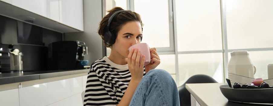 Close up portrait of smiling brunette woman, student drinks her tea and listens music or ebook in headphones, sits in kitchen and relaxes.