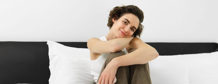 Close up portrait of young female model in white tank-top, sitting on bed in her bedroom and smiling at camera, leaning on pillows.