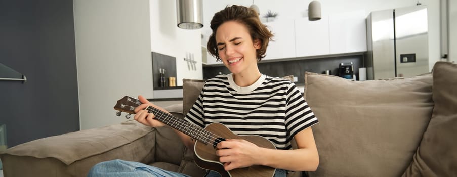 Portrait of young modern woman, student playing ukulele at home, sitting with small guitar, singing and feeling happy, sitting on sofa. Lifestyle and music concept