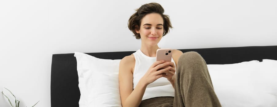 Portrait of young beautiful woman lying in bed, resting in bedroom, messaging, using mobile phone, holding smartphone and smiling, scrolling social media, spending comfort time at home.
