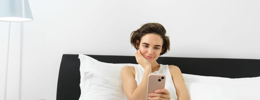 Lifestyle and technology concept. Smiling beautiful woman lying in bed with smartphone, resting in bedroom, chatting on social media, online shopping.