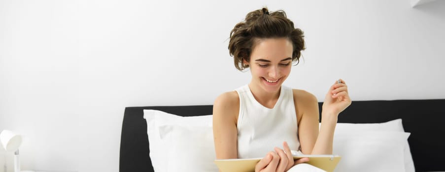Portrait of woman sitting in bed with journal, writing in diary with pen, reading notebook, studying or doing homework from her bedroom. Copy space