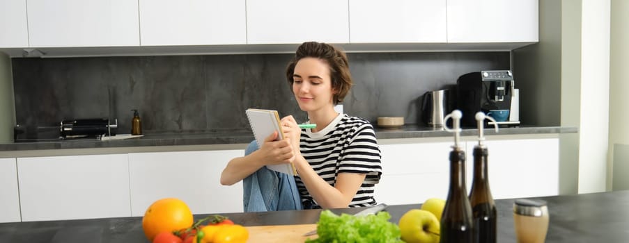 Portrait of beautiful modern woman, girl cooking with vegetables, holding notebook, writing down recipe, making notes, dinner plans, creating meal list for week, sitting in kitchen.