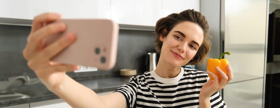 Cute and stylish lifestyle blogger, young woman taking selfie with fresh pepper, eating healthy vegetables, cooking salad in the kitchen and recording video on smartphone.