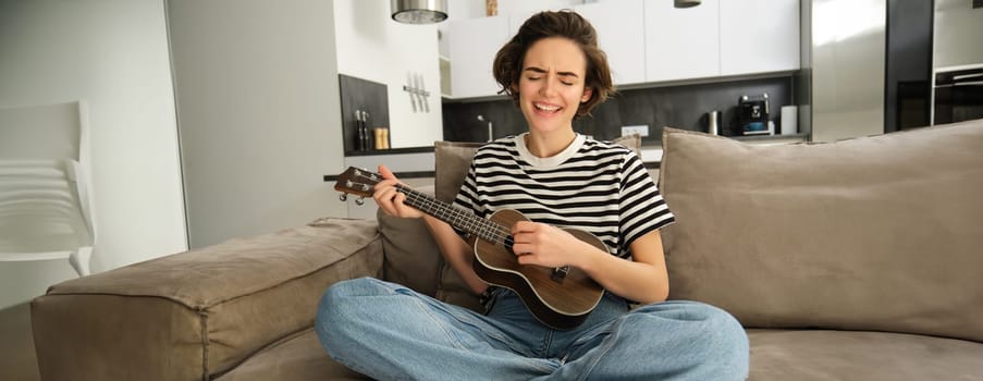 Portrait of cute young woman, girl with ukulele, playing and singing favourite song while sitting in living room on sofa, strumming strings.