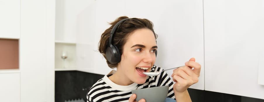 Happy beautiful woman, eating breakfast, cereals with milk as morning meal, holding bowl and spoon, listening music in wireless earphones, sitting in kitchen.