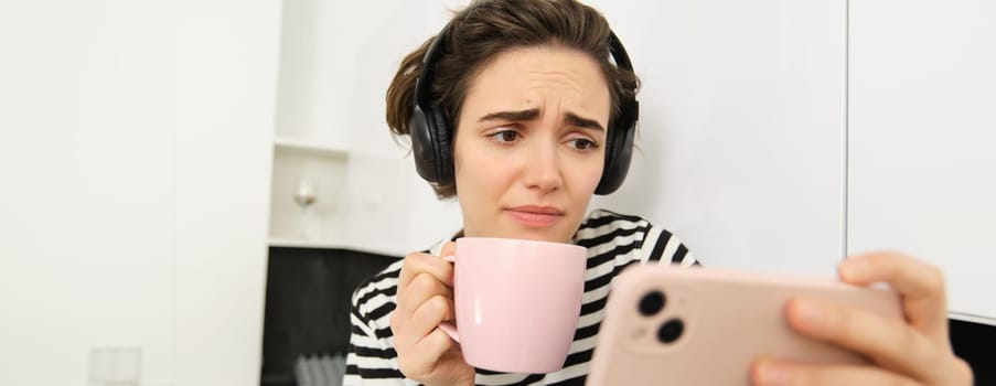 Portrait of woman with sad face, watching something on mobile phone, social media app, drinking tea, wearing wireless headphones.