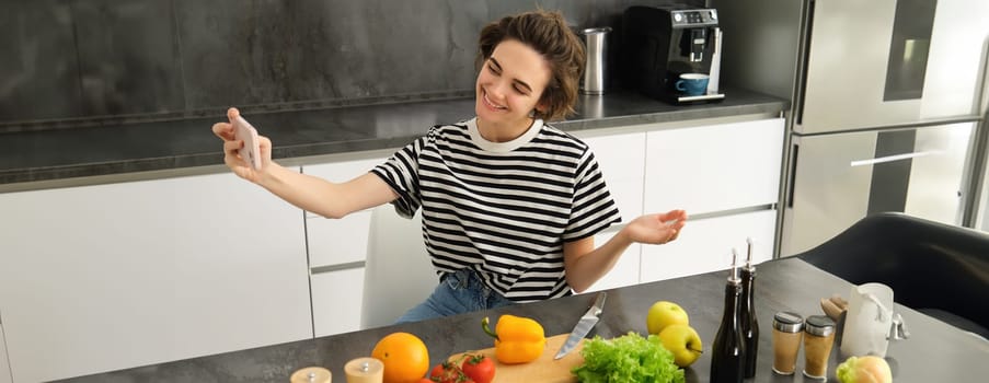 Portrait of beautiful young woman, food blogger recording video of her cooking in the kitchen, showing meal ingredients, preparing a meal on camera, using smartphone for live streaming.