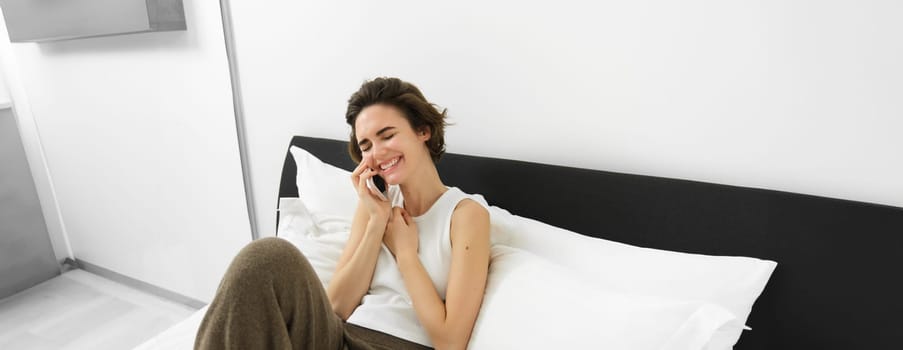 Female model sitting on bed with phone, calling friend, chatting over telephone, laughing and smiling.