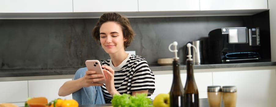 Portrait of young woman searching for cooking recipes online using smartphone, sitting near vegetables, salad ingredients and chopping board, smiling at camera.