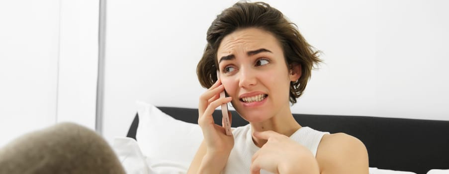 Image of woman with anxious, awkward face expression, talking on mobile phone, cringe from something she hears during conversation, lying in bed, saying something risky.