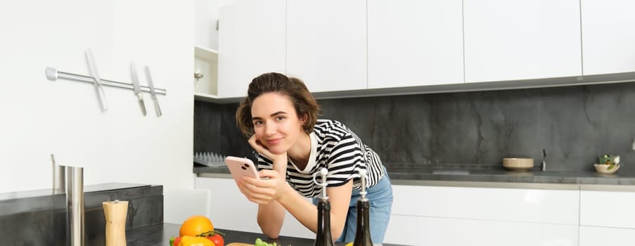 Woman bending on kitchen counter with smartphone, looking for healthy recipe online on mobile phone, standing next to chopping board and vegetables, cooking ingredients.