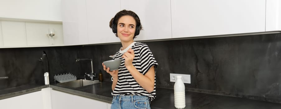 Portrait of modern woman eats breakfast, holds bowl and spoon, stands in kitchen, listens music in wireless headphones. Concept of lifestyle and home.