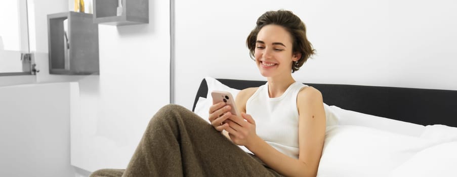 Portrait of young beautiful woman lying in bed, resting in bedroom, messaging, using mobile phone, holding smartphone and smiling, scrolling social media, spending comfort time at home.